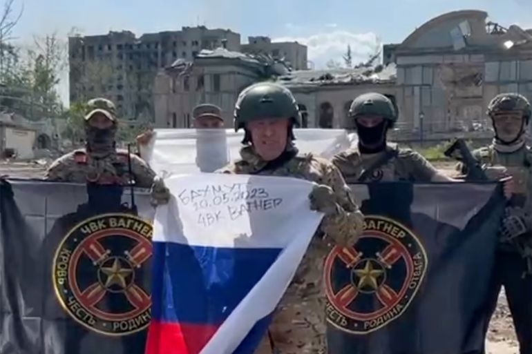Yevgeny Prigozhin pictured holding a Russian flag in ruined Bakhmut. He is in combat fatigues and there is group of soldiers standing behind him. There are destroyed buildings in the background