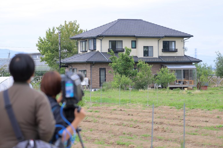 View of the two-story house where the suspect hid from the police.  The news crew of the television company is filming the building.  There is a field ahead.