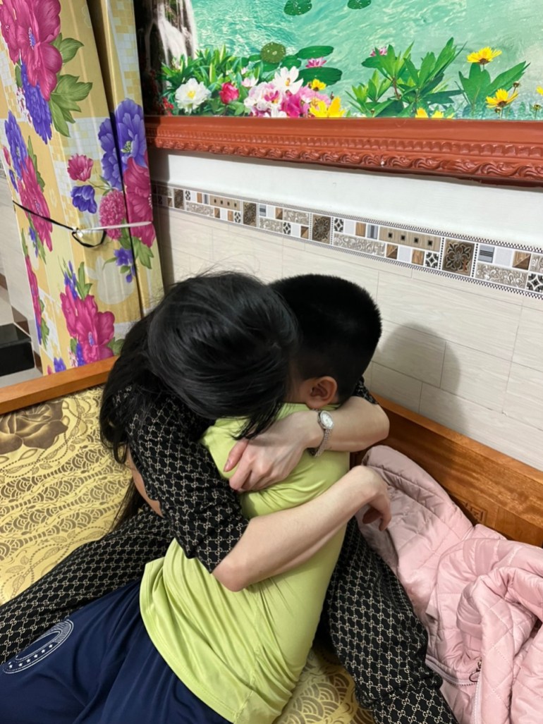 A woman hugging her son. Their faces are obscured. There is a colourful floral picture behind them.