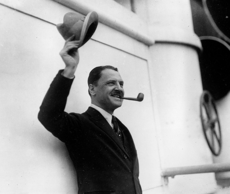 The writer W Somerset Maugham on the deck of a ship. He is holding his hat aloft and has a pipe in his mouth.