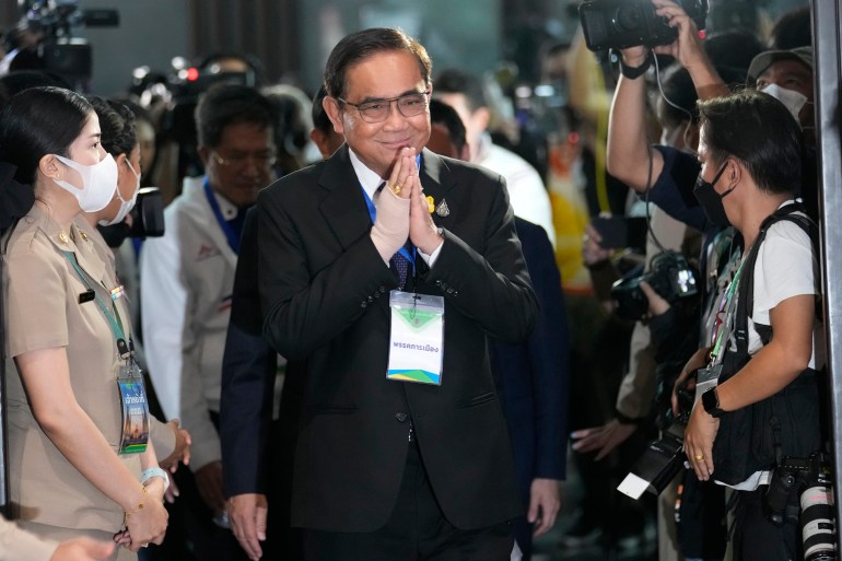 Prayuth Chan-ocha with his hands together in a traditional wai greeting. He is wearing a dark suit and smiling