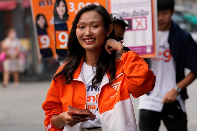 Chonticha Jungangru smiles and holds her left hand in her hair while others clutch a booklet. She wears a jacket with orange on her top and her sleeves and white on her lower half. Unzipping her reveals a white T-shirt with orange lettering.
