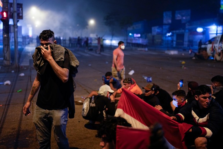 A man covers his face with fabric, while other protesters nearby hold a flag and sit on the ground