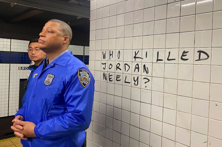 A police officer in a blue shirt stands in front of a white tiled wall with the inscription: Who killed Jordan Neely?
