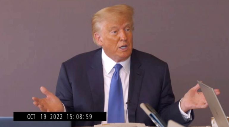 Donald Trump, in a suit and blue tie, speaks into a microphone against a mauve background. A time stamp in the bottom of the picture indicates this is a video still