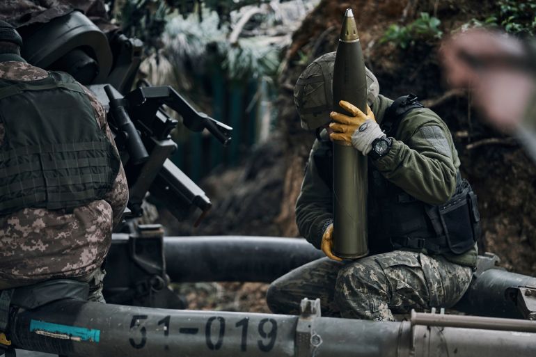 Um soldado ucraniano, à direita, está sentado e segurando um projétil de artilharia [Libkos via AP]