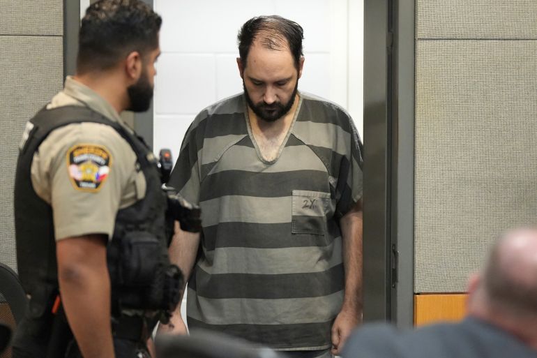 A man in a grey-and-black striped jumpsuit enters a court room, escorted by guards