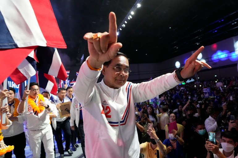 Prayuth memberi isyarat kepada para pendukungnya.  Dia mengenakan baju olahraga dengan warna pesta merah putih dan biru dan ada bendera di latar belakang.