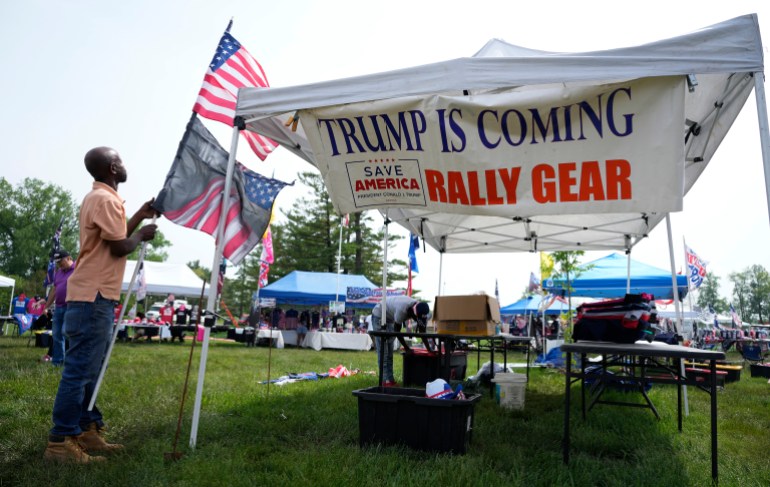 A man disassembles a stack of flags, next to a white tent with a banner that reads: "Trump is coming. Rally gear."