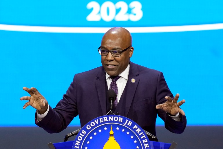 Kwame Raoul, wearing a blue suit, gestures with both hands behind a podium. He is standing in front of a backdrop that reads "2023."
