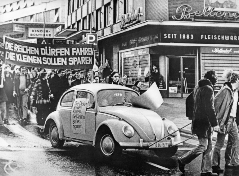 A Volkswagen Beetle towed though an Essen street in Germany as part of a protest agains a driving ban imposed during the oil crisis in 1973. Protesters are walking behind with a large banner. The photo is in black and white.