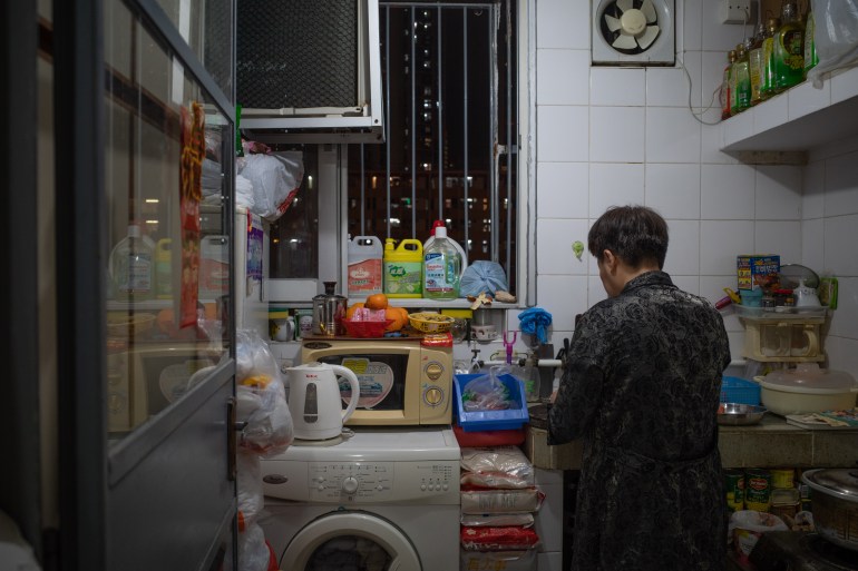 السيدة تشان في المطبخ الضيق في شقة عائلتها المستأجرة والتي تبلغ مساحتها 23 مترًا مربعًا (248 قدمًا مربعًا).  يمكن رؤية المواد الغذائية ومنتجات التنظيف مرتبة في كل زاوية وركن متاح. 