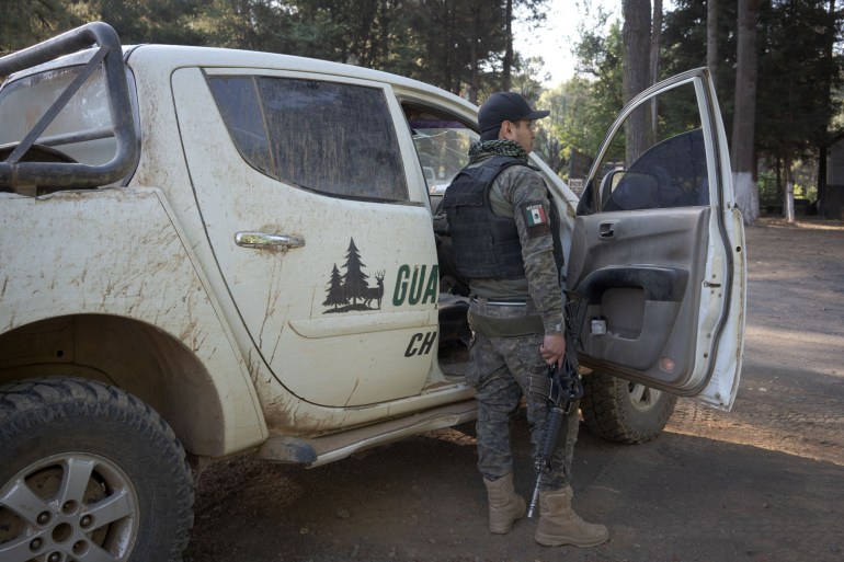 مردی با لباس نظامی و جلیقه ضد گلوله در مقابل یک کامیون سفید ایستاده است.
