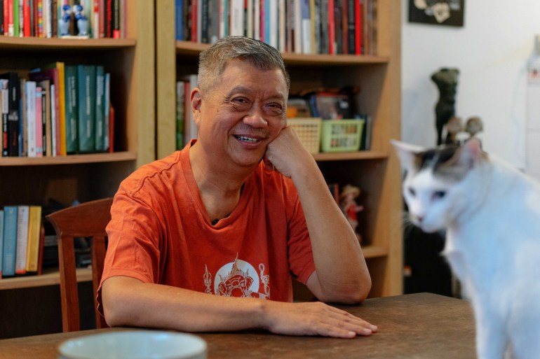 Dédé Oetomo sitting at a desk with a book case behind him. He is smiling and propping his head up on one hand. There's a calico cat walking on the desk in front.