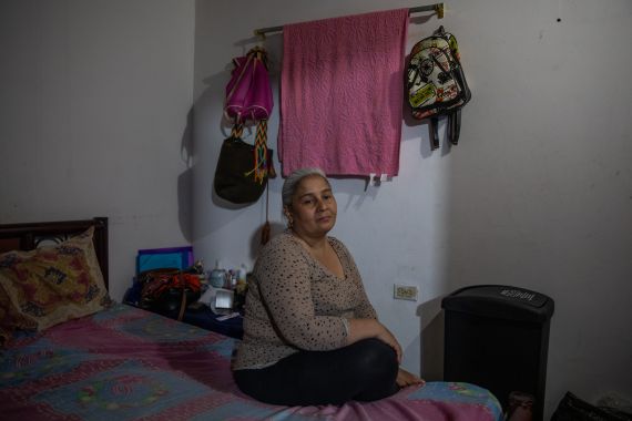 A woman sits on a bed in a darkened apartment, with a pink blanket and book bags hanging behind her