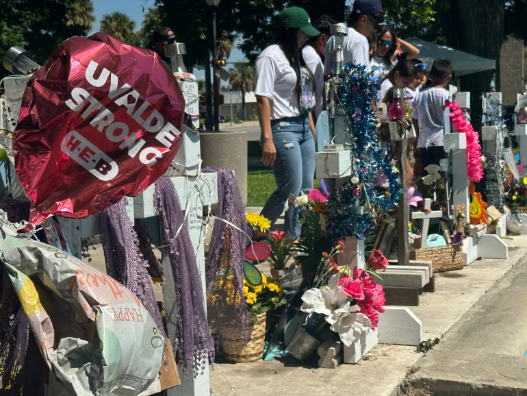 Cư dân Uvalde đang bày tỏ lòng kính trọng đối với những đứa trẻ và giáo viên đã bị bắn chết tại Trường Tiểu học Robb một năm trước.  Có những bó hoa, thánh giá và bóng bay.  Quả bóng lớn màu đỏ ở phía trước ghi 'Uvalde Strong' 