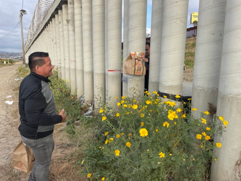 Between pillars of concrete, a hand holds out a bag of food to an asylum seeker