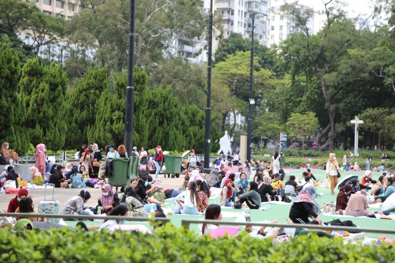 Pekerja rumah tangga Indonesia nongkrong di Victoria Park pada hari libur mereka.  Mereka duduk di tanah mereka.  Ada pepohonan dan gedung pencakar langit di belakang mereka.