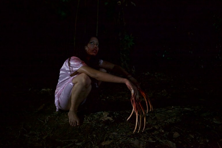 A still from the movie Lembu showing a young woman squatting down in the darkness. She is wearing a pink dress and barefoot. Her fingers are elongated and covered in blood.