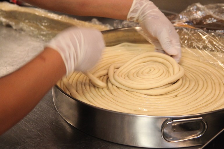 A circular metal pan holds a long spiral of noodles. A pair of gloved hands gently readjusts the latest layer of noodle.