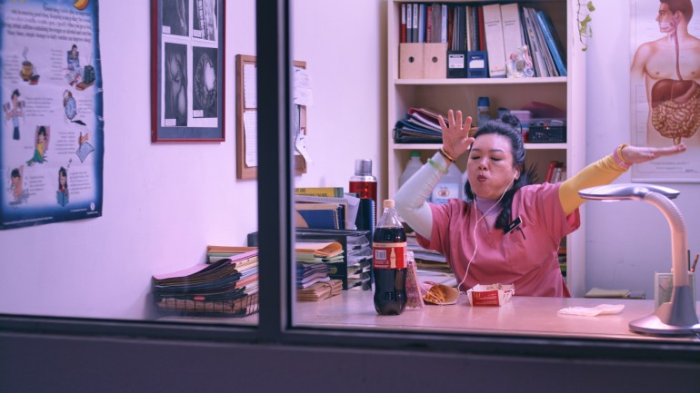 A still from Vinegar Baths shows a woman sitting at a desk in what looks like a clinic. Anatomical charts hung on the walls, and behind her was a bookcase with papers. There is a bottle of coke and some french fries on her table. She wears headphones and moves her arms over the music playing on her phone.