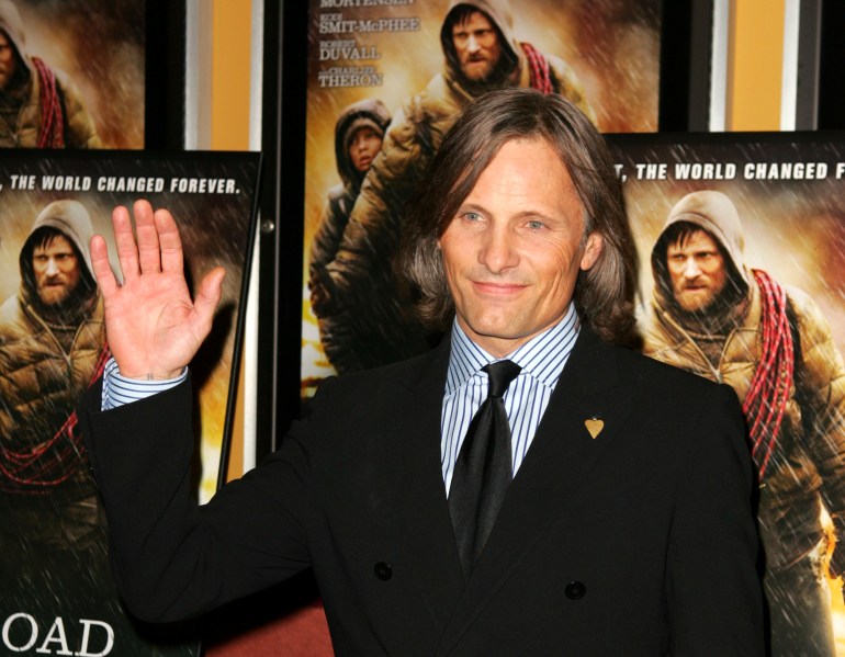 A man in a dark suit waves in front of a movie poster of The Road.