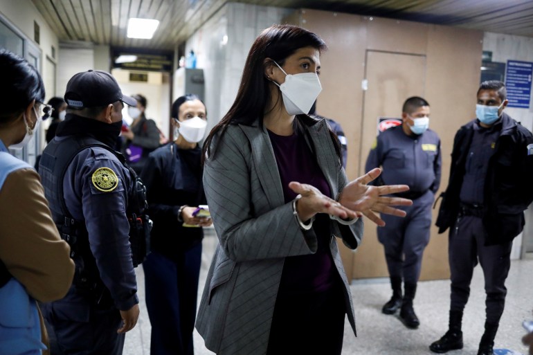 A lawyer in a grey suit and black shirt gestures in handcuffs, as police follow her. They all wear COVID face masks.
