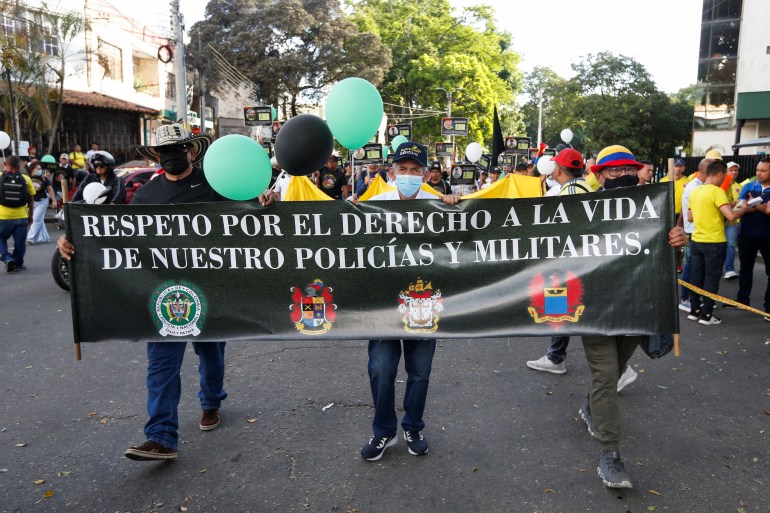 Three men on the streets of Colombia hold up a black banner with white text, that displays police and military seals. Balloons can be seen behind them.