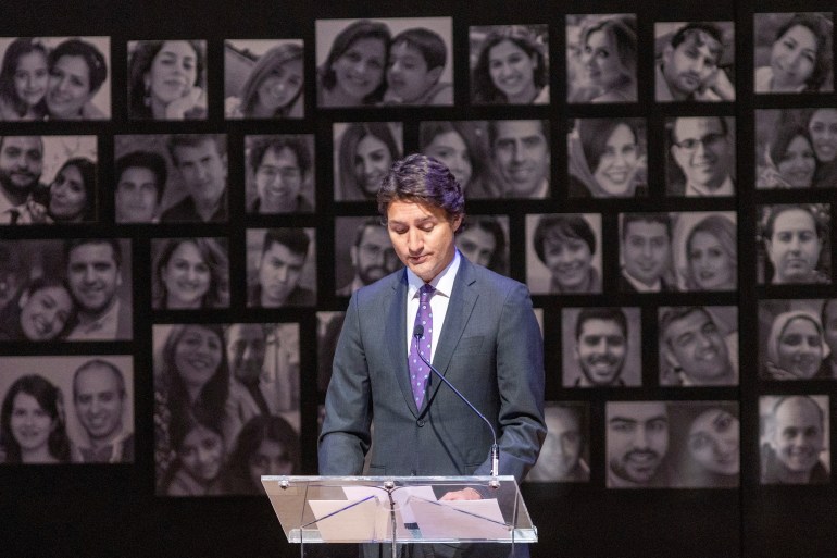 Prime Minister Justin Trudeau — in a grey suit and purple tie — stands at a glass podium. Behind him is a wall of black-and-white photos depicting the lives lost in the plane crash.