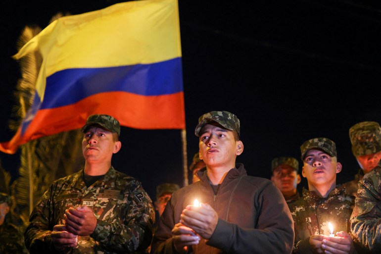 Soldiers against a dark night sky, dressed in military fatigues, hold candles and a large Colombia flag.