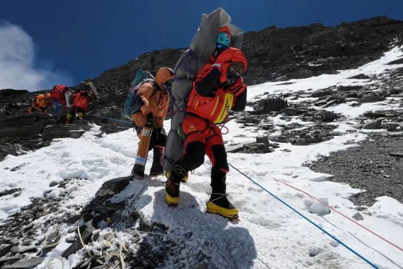 Ngima Tashi Sherpa walks with a Malaysian climber on his back from Mount Everest. It is snowy and rocks are visible. The climber is wrapped in a sleeping mat. Another sherpa is behind.