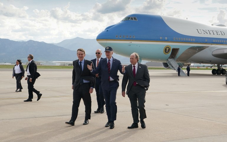 With Air Force One plane behind him, President Joe Biden walks across the tarmac, surrounded by men in suits.