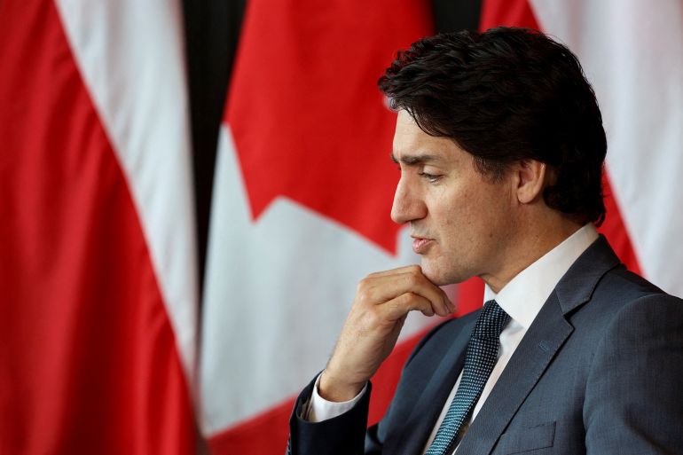 Justin Trudeau, dressed in suit and tie, leans his head on his hand, against a backdrop of Canadian flags.