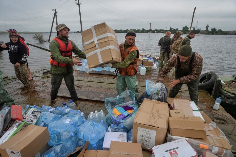 Ukrainian soldiers helping with flood relief efforts after the Nova Kakhovka dam breach. Theye are moving large boxes of supplies. There are more boxes and bags in front of them.