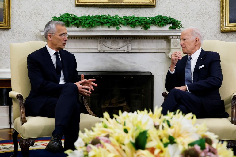Two men in dark suits sit in the Oval Office of the White House. A fire place is visible behind them, and flowers on a table before them.