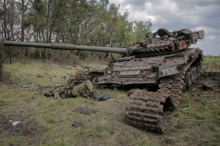 A destroyed Russian tank. The body of a soldier lies in front of it. Their face cannot be seen.