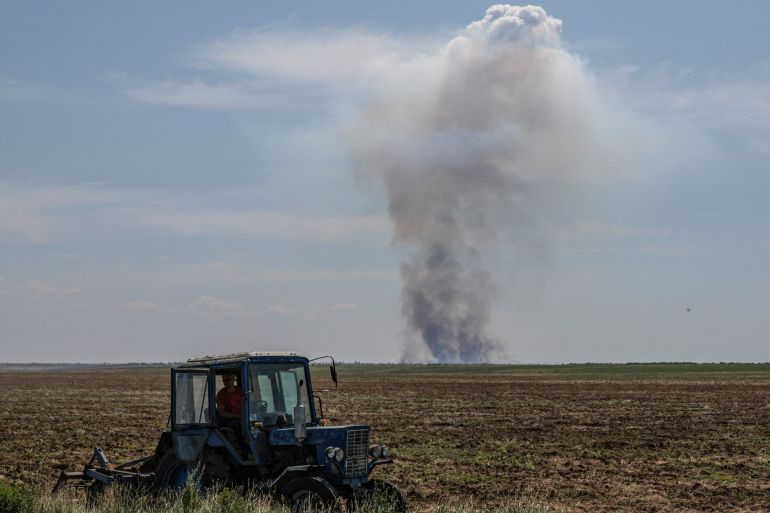A farmer driving a tractor in a vast field. A column of smoke is rising on the horizon after a military attack