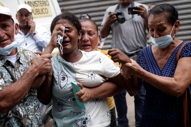 A woman in a white T-shirt holds a paper face mask over her eyes as she cries, surrounded by other bereaved relatives.