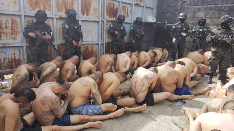 Suspected gang members sit closely packed in lines on the ground, stripped naked except for boxer shorts, as armed guards watch over them from near a prison wall.