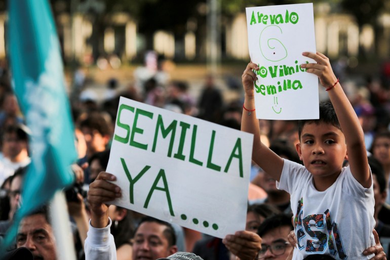 People outdoors gather, holding up hand-written signs reading, "Semilla ya." One child sits atop an adult's shoulders.