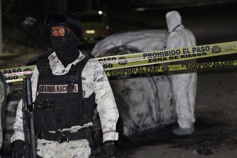 A man in a black flak jacket, white camouflage and a face mask walks away from a burnt-out car that is being processed as a crime scene, surrounded by yellow caution tape.