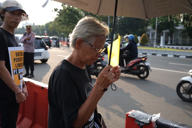 Maria Catarina Sumarsih. The 71-year-old is at a rally outside the Presidential Palace over Indonesian rights abuses. She has white hair and is wearing glasses. She is standing beneath an umbrella. Another protester is beside her.