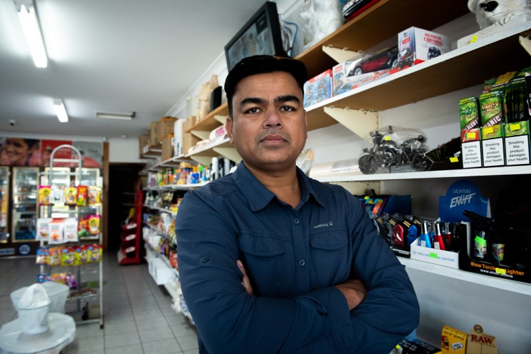 Habiburahman, a Rohingya in exile. He is in his stop and standing with his arms folded. He is wearing a blue shirt. There are shelves stacked with goods behind him.