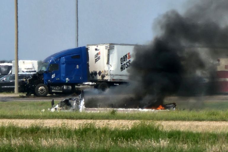Black smoke billowing from a burning vehicle after a crash in Manitoba. There is a large truck behind with its engine smashed.