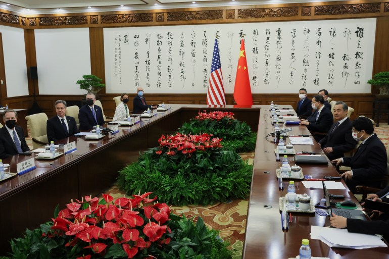 American and Chinese delegations at the table.  They are sitting across from each other at a long table.  There are plants in the middle.  The flags of the countries and Chinese calligraphy follow.