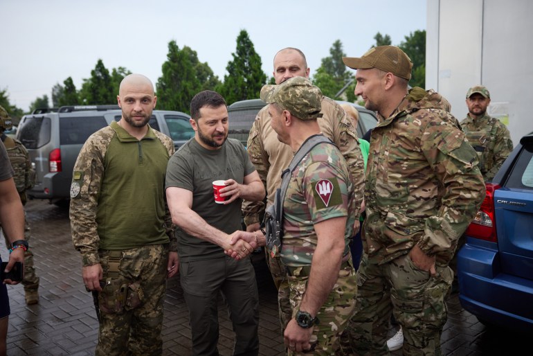 Volodymyr Zelenskyy, Donetsk'te ön cephe yakınında Ukraynalı askerlerle el sıkışırken.  Haki renkli bir tişört giyiyor ve sol elinde bir paket içecek tutuyor.