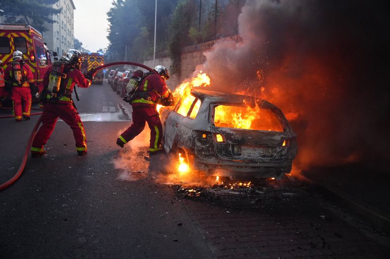 İtfaiyeciler, polisin trafik kontrolü için durmayı reddeden bir genci öldürmesinin ardından 27 Haziran 2023'te Paris'in batısındaki Nanterre'deki bir gösterinin kenarlarında yanan bir arabayı söndürmek için çalışıyor