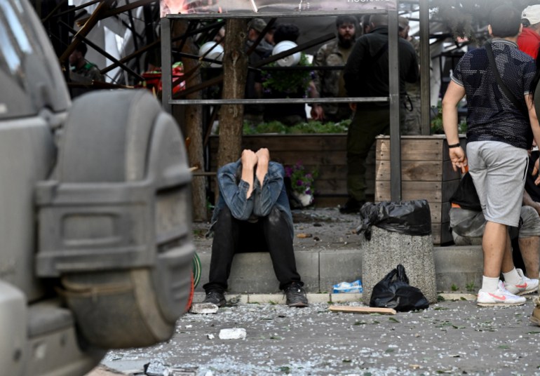A man covers his face with his hands outside the destroyed restaurant. He is sitting on the side of the road. There is debris around and in front of him. He looks in despair.