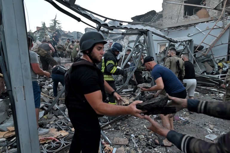 People work to rescue those caught in the missile attack on Kramatorsk. There is a twisted metal frame above them and people working to shift the rubble beneath. In the front of the photo, a man is passing debris to another person in the chain.