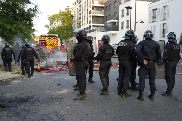 Police in riot gear stand guard as firefighters work to put out fires after a demonstration in Nanterre, west of Paris, on June 27, 2023, after French police killed a teenager who refused to stop for a traffic check in the city. - The 17-year-old was in the Paris suburb early on June 27 when police shot him dead after he broke road rules and failed to stop, prosecutors said. The event has prompted expressions of shock and questions over the readiness of security forces to pull the trigger. (Photo by Zakaria ABDELKAFI / AFP)
